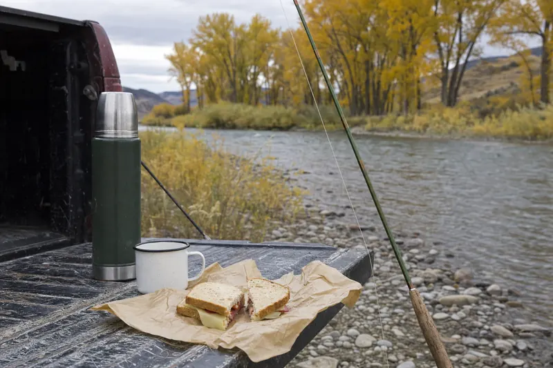 A riverside lunch on a pickup tailgate: wrapped sandwiches, a thermos and an enamel mug.